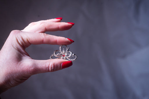 May include: A silver tiara with a red gem in the center. The tiara has a delicate, ornate design with small, sparkling stones.