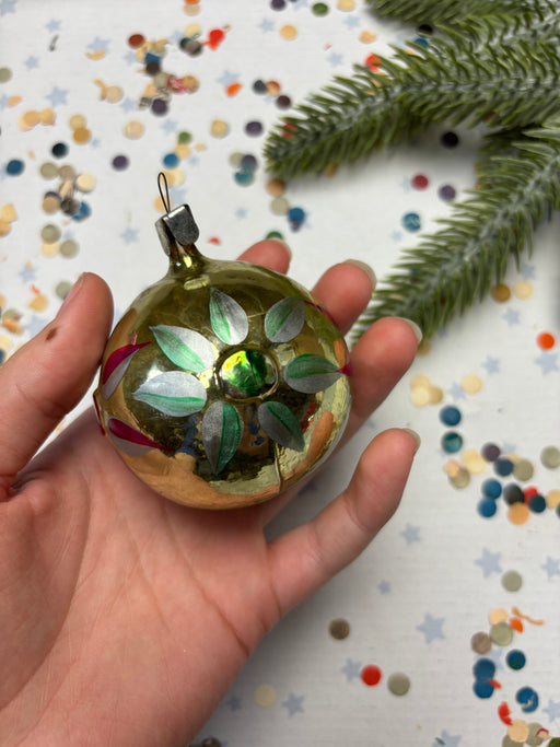 A hand holding a gold ornament with green leaves and a small green gemstone, surrounded by colorful confetti on a white background.