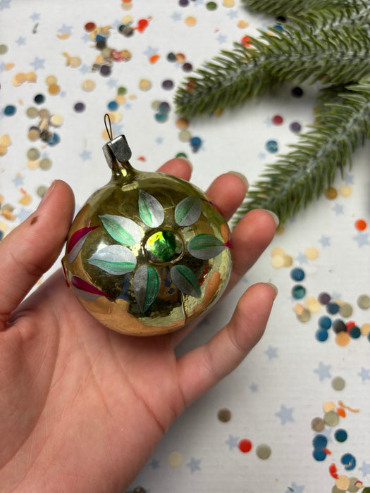 A hand holding a gold ornament with green leaves and a small green gemstone, surrounded by colorful confetti on a white background.