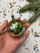A hand holding a gold ornament with green leaves and a small green gemstone, surrounded by colorful confetti on a white background.