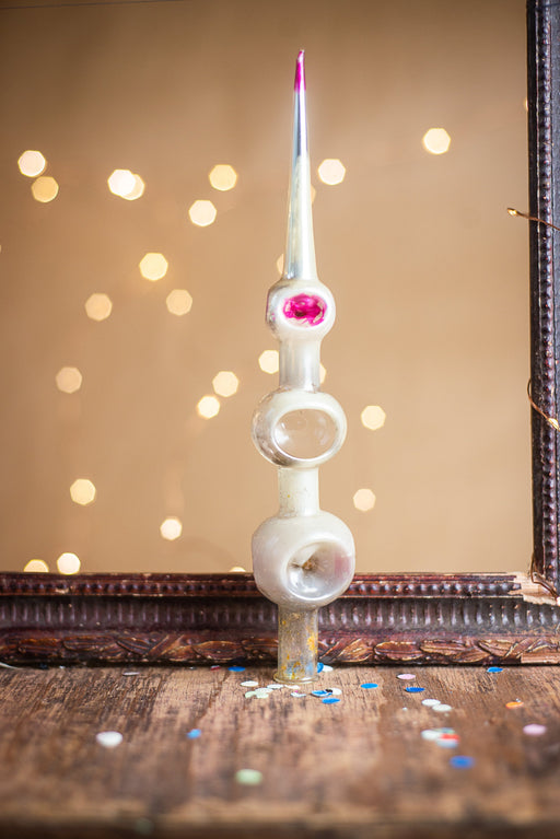 A decorative glass ornament with a pointed top and three circular openings is displayed on a wooden surface, surrounded by scattered confetti.