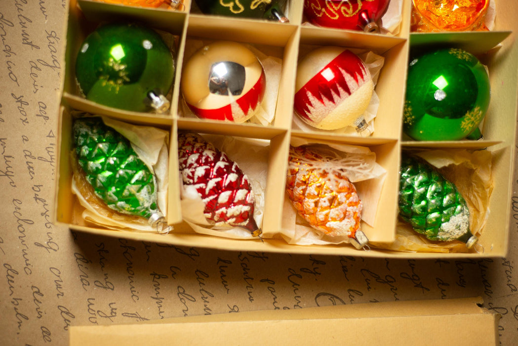 A cardboard box containing a variety of Christmas ornaments, including green, red, and orange balls, as well as pine cone-shaped ornaments.