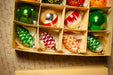 A cardboard box containing a variety of Christmas ornaments, including green, red, and orange balls, as well as pine cone-shaped ornaments.