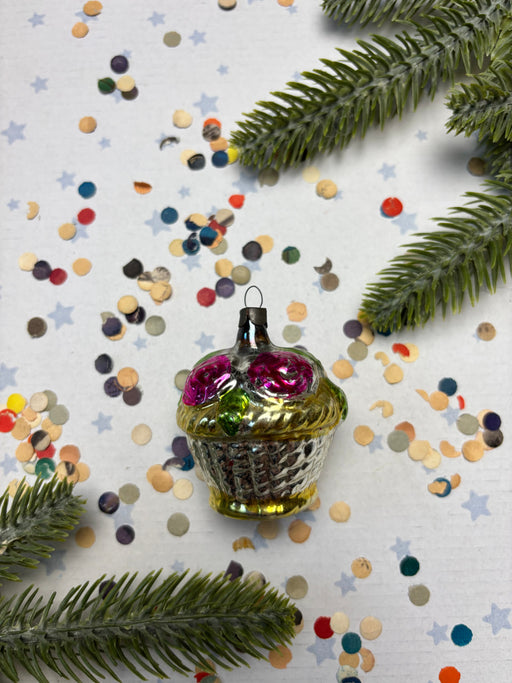 A glass ornament with a gold base and pink flowers sits on a white background, surrounded by colorful confetti and pine branches.