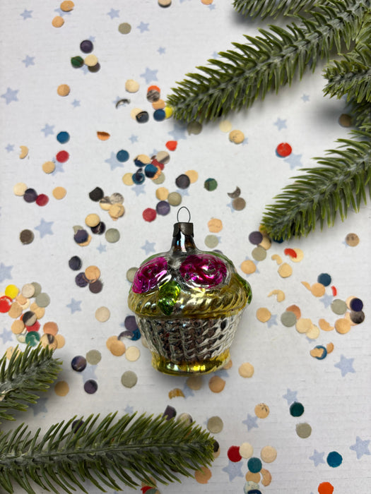 A glass ornament with a gold base and pink flowers sits on a white background, surrounded by colorful confetti and pine branches.
