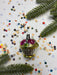 A glass ornament with a gold base and pink flowers sits on a white background, surrounded by colorful confetti and pine branches.