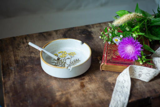A white ceramic ashtray with a floral design sits on a wooden surface next to a book and a purple flower.