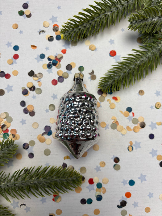 A silver ornament with a pointed top is surrounded by colorful confetti and green pine branches on a white background.