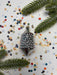 A silver ornament with a pointed top is surrounded by colorful confetti and green pine branches on a white background.