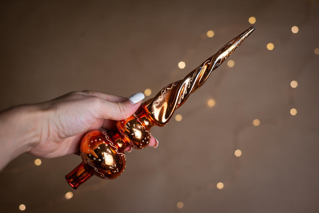 A hand holding a decorative Christmas ornament with a twisted, metallic design against a blurred background with twinkling lights.