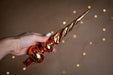 A hand holding a decorative Christmas ornament with a twisted, metallic design against a blurred background with twinkling lights.