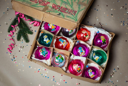 A cardboard box filled with colorful Christmas ornaments, including Smurfs, is surrounded by confetti on a table.