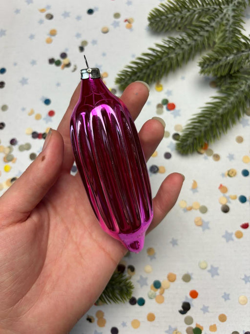 A hand holding a pink ornament against a background of confetti and pine branches.
