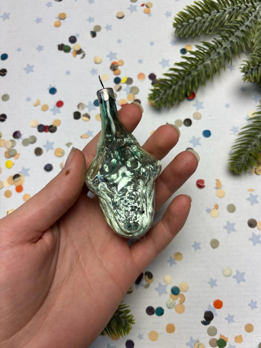 A hand holding a green glass ornament with a silver clasp, surrounded by colorful confetti and a sprig of greenery.
