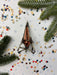 a metallic ornament with a triangular shape, surrounded by colorful confetti and pine branches.