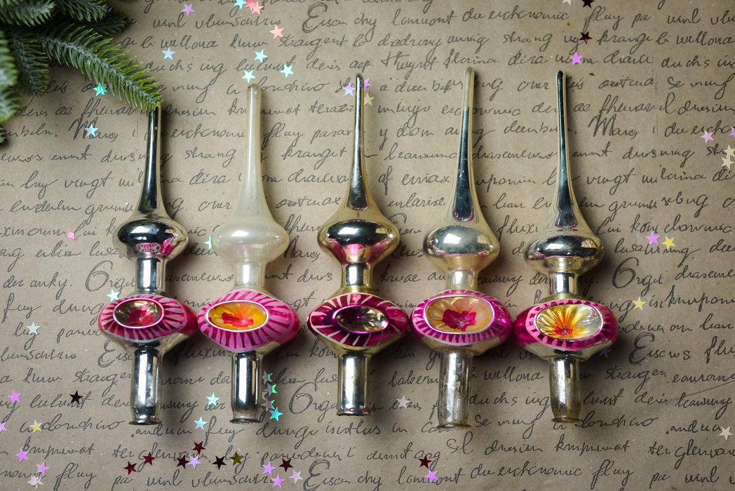 A row of six Christmas tree ornaments with pink and yellow flowers, arranged on a piece of paper with handwritten text.