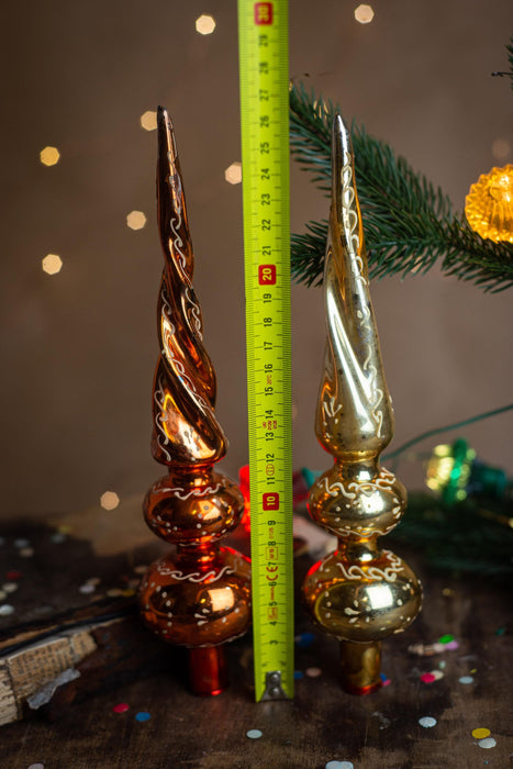 two ornate Christmas tree ornaments, one in gold and the other in copper, placed next to a green pine branch with twinkling lights.