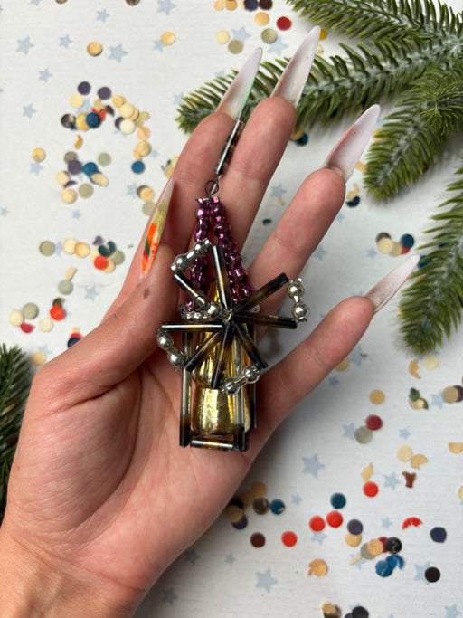 A hand holding a decorative piece with metallic and colorful elements, surrounded by confetti and pine branches.