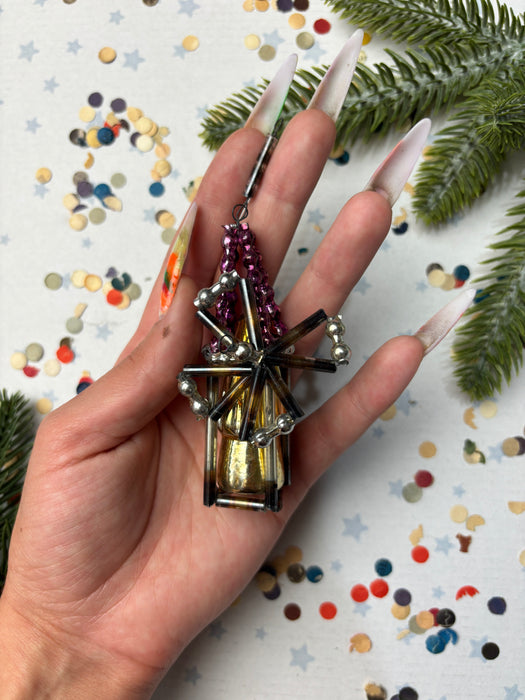 A hand holding a decorative piece with metallic and colorful elements, surrounded by confetti and pine branches.