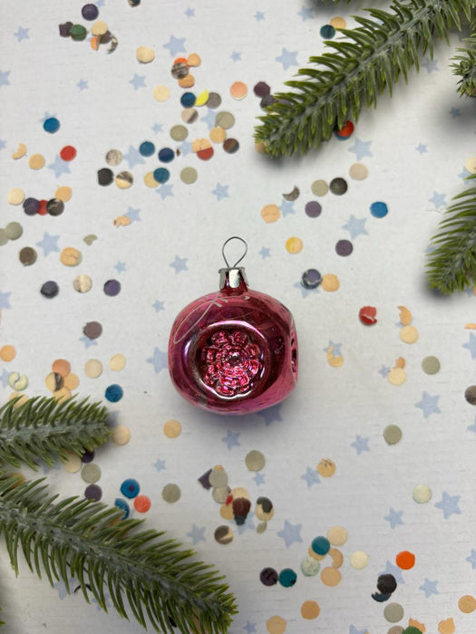 A red Christmas ornament with a shiny surface sits on a white background, surrounded by scattered confetti and green pine branches.