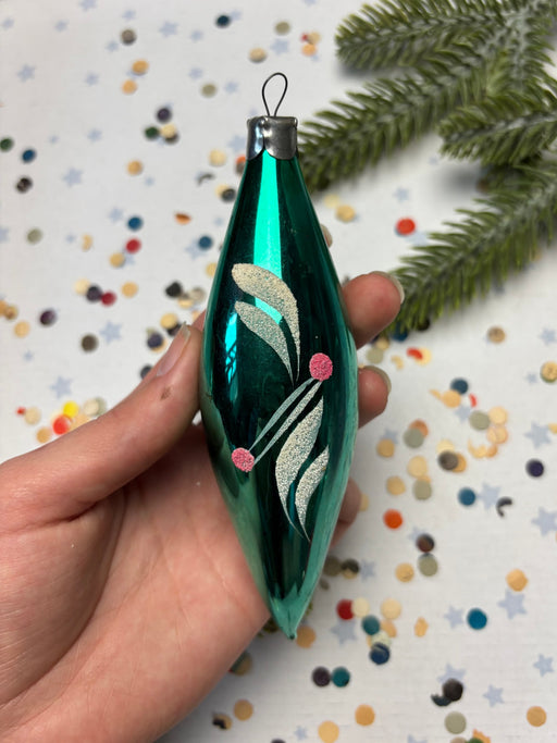 A hand is holding a green glass ornament with a floral design, surrounded by colorful confetti on a white background.