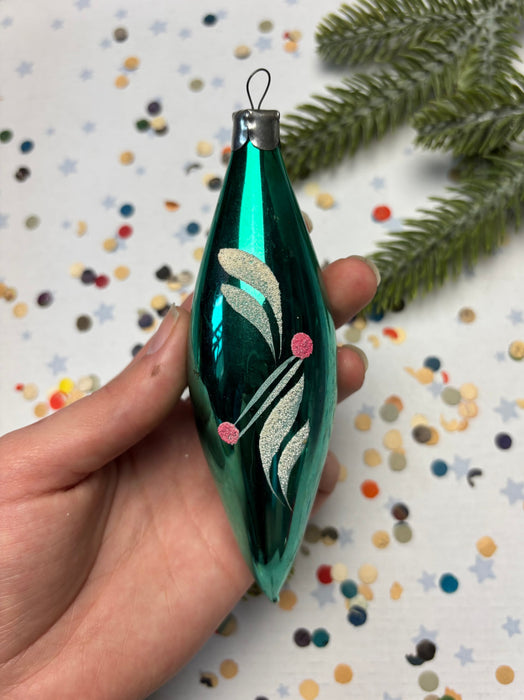 A hand is holding a green glass ornament with a floral design, surrounded by colorful confetti on a white background.