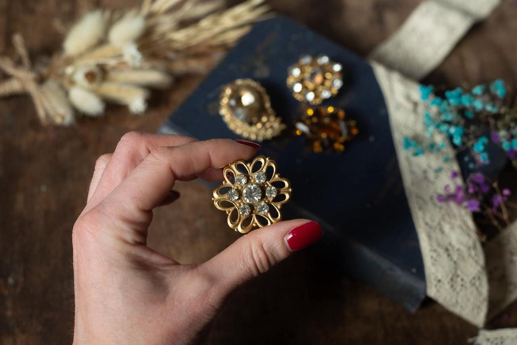 A hand holding a gold brooch with a floral design, surrounded by various jewelry pieces and flowers on a wooden surface.