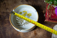A white ceramic ashtray with a floral design sits on a wooden surface, with a yellow measuring tape extending across it.