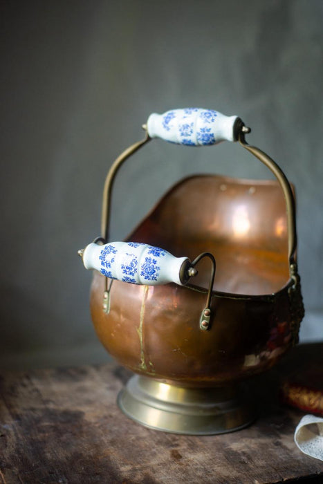 A copper-colored metal bowl with a handle and two blue floral handles, placed on a wooden surface.