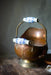 A copper-colored metal bowl with a handle and two blue floral handles, placed on a wooden surface.