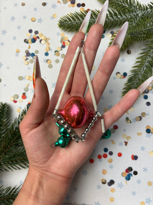 A hand holding a Christmas ornament with a red and green color scheme, surrounded by pine branches and confetti.