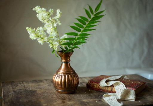 A copper vase holding white flowers and green leaves sits on a wooden table, accompanied by a red book with a white lace ribbon on top.