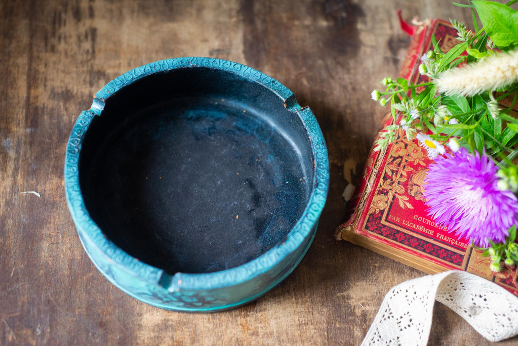 An empty blue ashtray sits next to a red book with a purple flower on the cover, placed on a wooden surface.