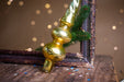 A golden ornament with a curved top and two round bases is leaning against a dark wooden surface, with a sprig of greenery and scattered confetti nearby.