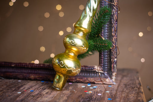 A golden ornament with a curved top and two round bases is leaning against a dark wooden surface, with a sprig of greenery and scattered confetti nearby.