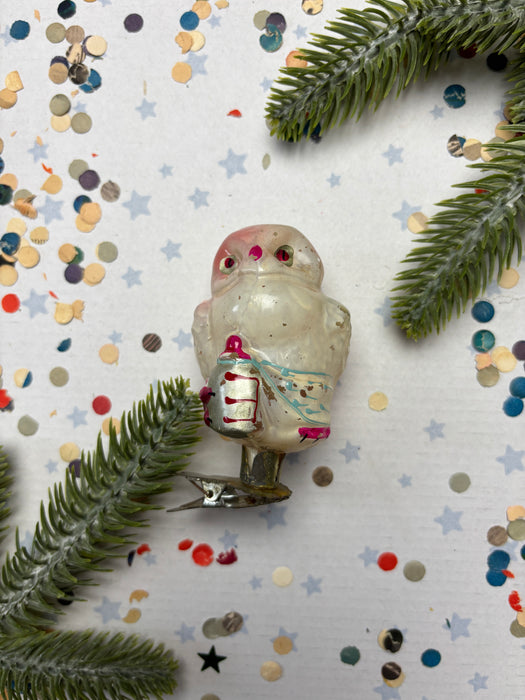 a small, white owl ornament with a red bow around its neck, placed on a white background with scattered confetti and pine branches.