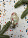 a small, white owl ornament with a red bow around its neck, placed on a white background with scattered confetti and pine branches.