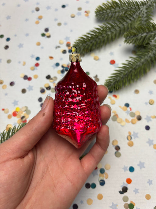 A hand holding a red glass ornament with a gold-colored stem, surrounded by scattered confetti and a sprig of greenery.