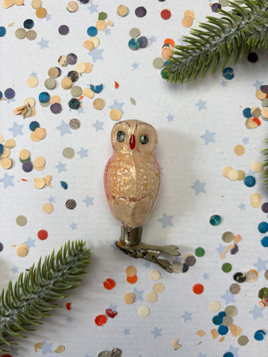 a small, beige owl ornament placed on a white background, surrounded by colorful confetti and green pine branches.