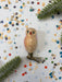 a small, beige owl ornament placed on a white background, surrounded by colorful confetti and green pine branches.