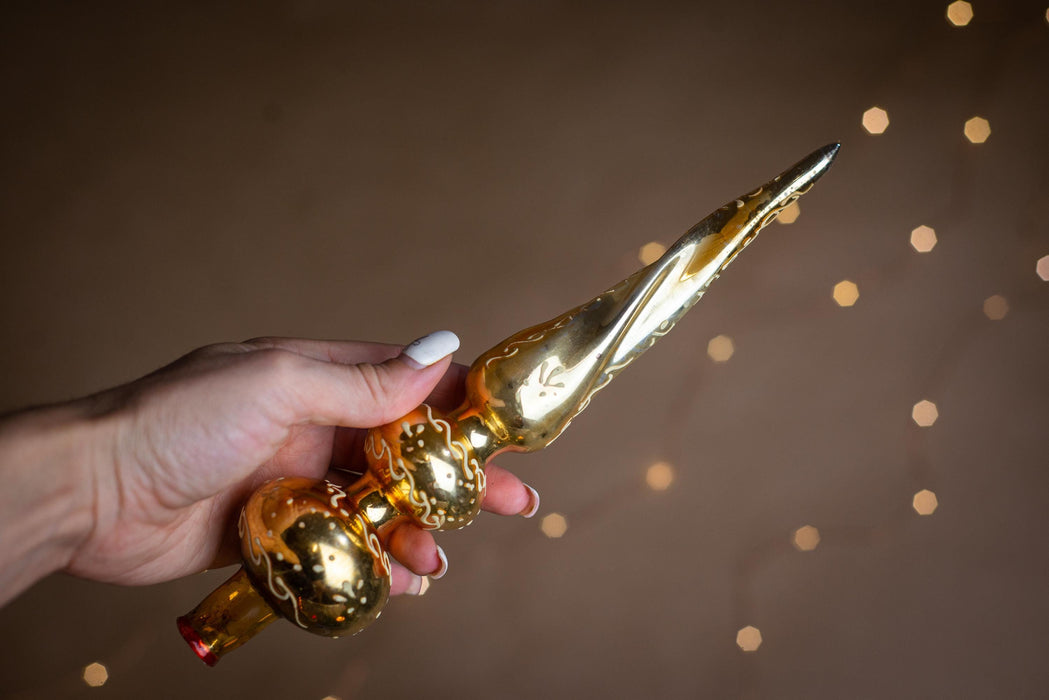 A hand holding a golden ornament with a pointed end, against a background of blurred lights.