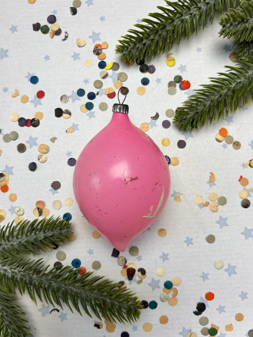 A pink Christmas ornament is surrounded by confetti and pine branches on a white background.
