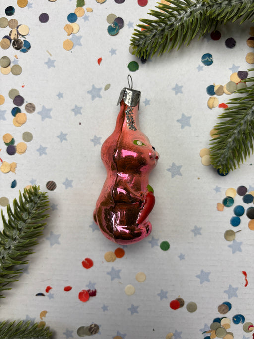 A pink glass ornament in the shape of a cat is surrounded by confetti and pine branches on a white background.