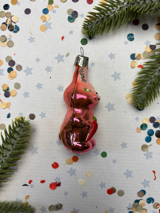 A pink glass ornament in the shape of a cat is surrounded by confetti and pine branches on a white background.