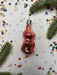 A pink glass ornament in the shape of a cat is surrounded by confetti and pine branches on a white background.
