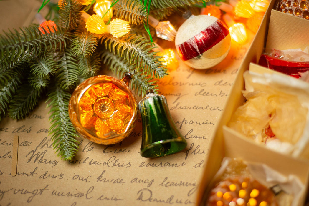 a close-up view of a Christmas tree adorned with ornaments, including a bell, a ball, and a star, all surrounded by twinkling lights. The ornaments are placed on a piece of paper with handwritten text, creating a festive and nostalgic atmosphere.