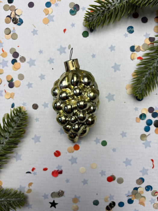 A shiny gold ornament with a bunch of small balls is displayed against a background of colorful confetti and pine branches.