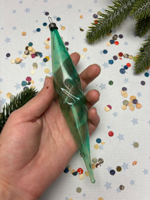A hand is holding a green glass ornament with a twisted shape, surrounded by colorful confetti and pine branches.