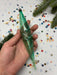 A hand is holding a green glass ornament with a twisted shape, surrounded by colorful confetti and pine branches.