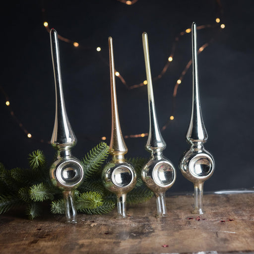 four ornate Christmas ornaments with pointed tops, arranged on a wooden surface. The ornaments are silver, gold, and bronze in color, and they are set against a dark background with twinkling lights.